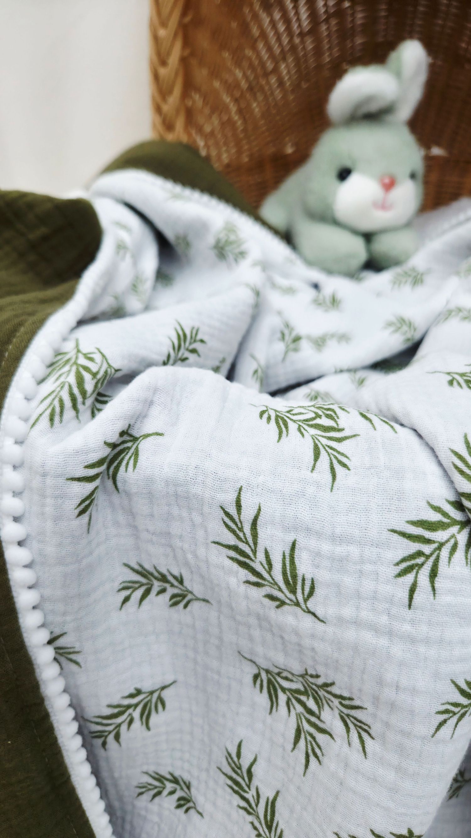 Light blue muslin baby blanket with green leafy sprig print and white pom-pom trim, with a plush gray-green bunny toy in a wicker basket.
