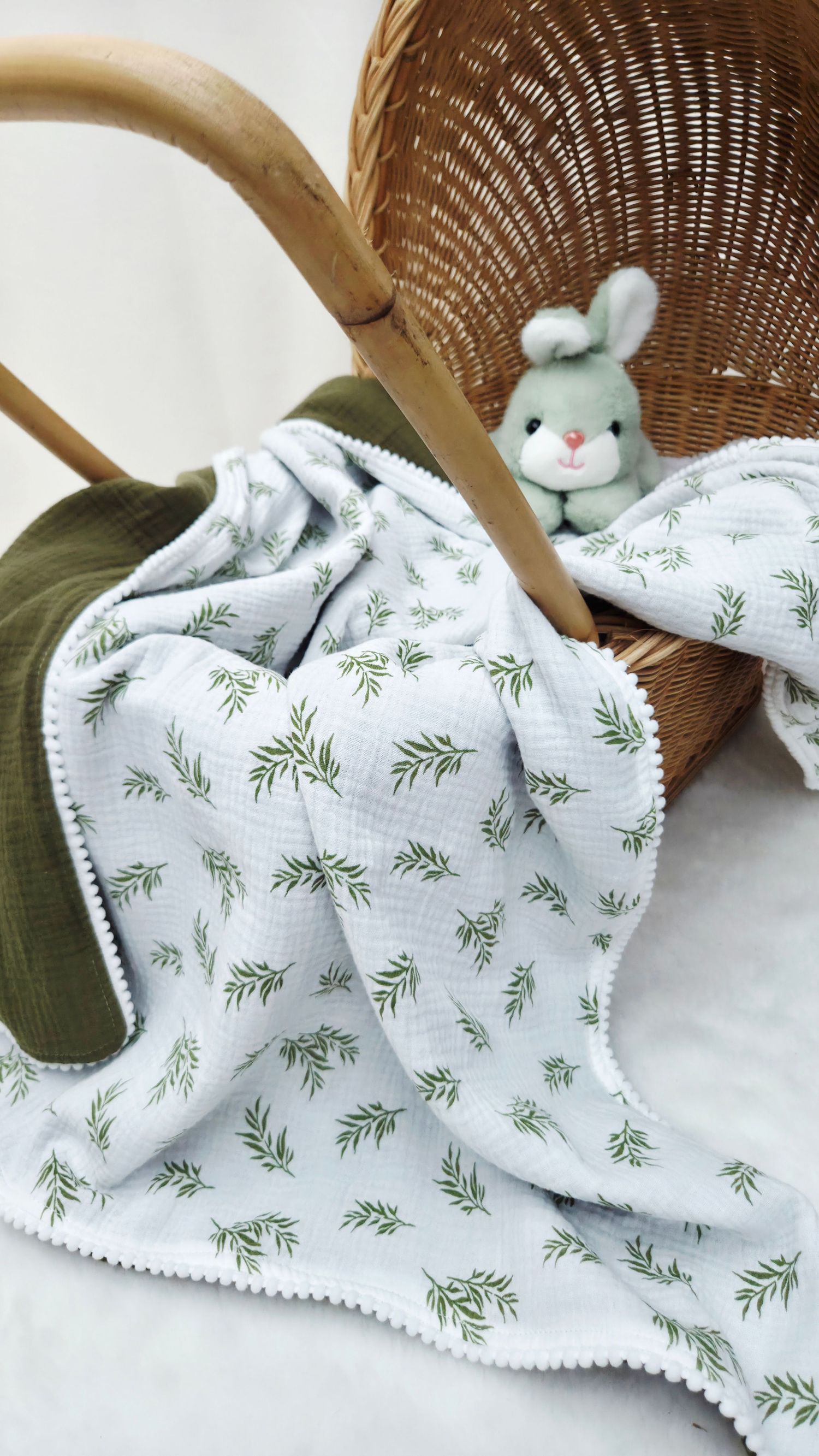 Soft muslin baby blanket with green leaf print and pom-pom trim draped over a wicker basket beside a plush sage green bunny toy.