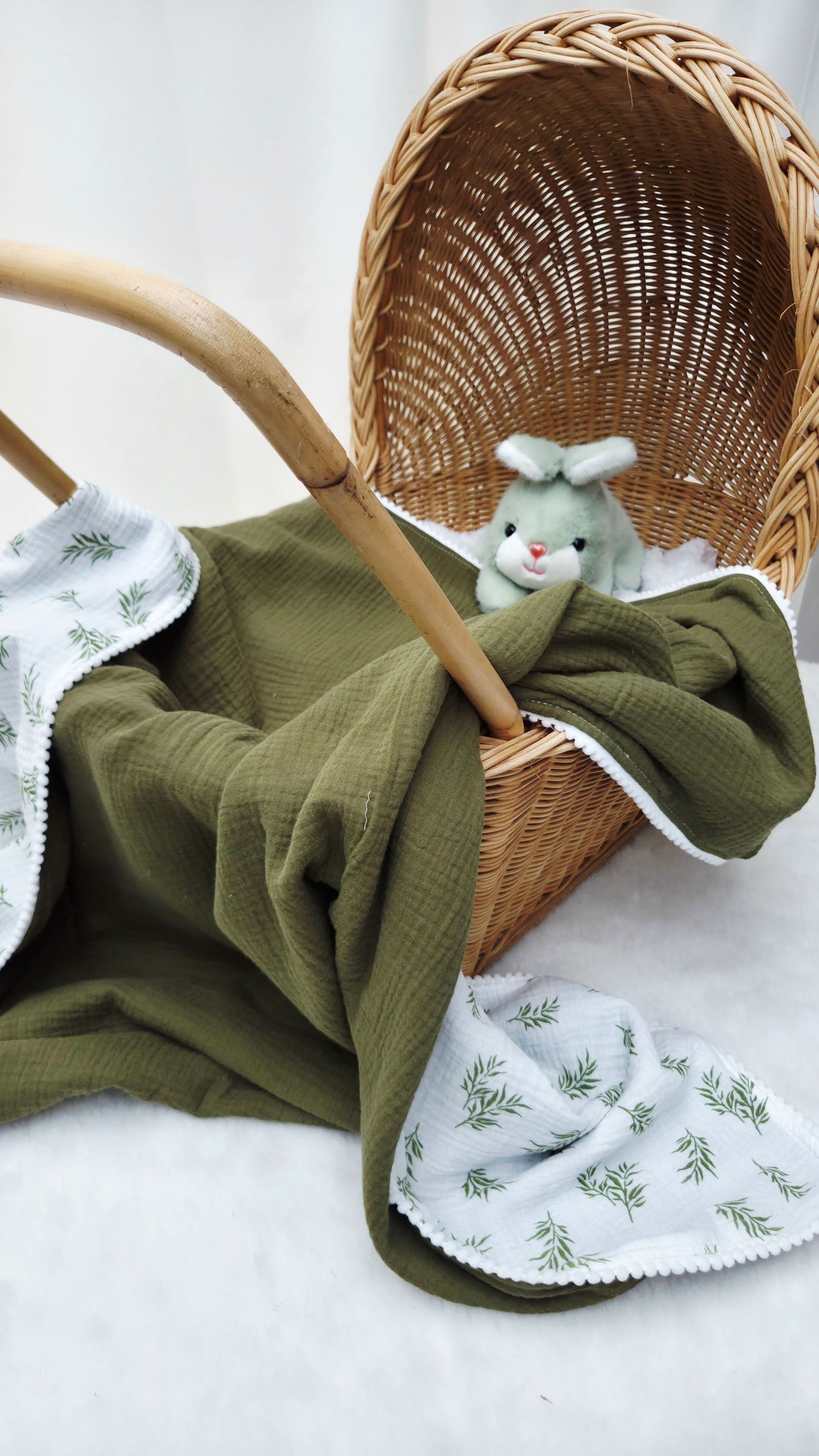 Olive green muslin baby blanket with white leaf-print trim draped over a wicker bassinet basket holding a soft sage green bunny toy.