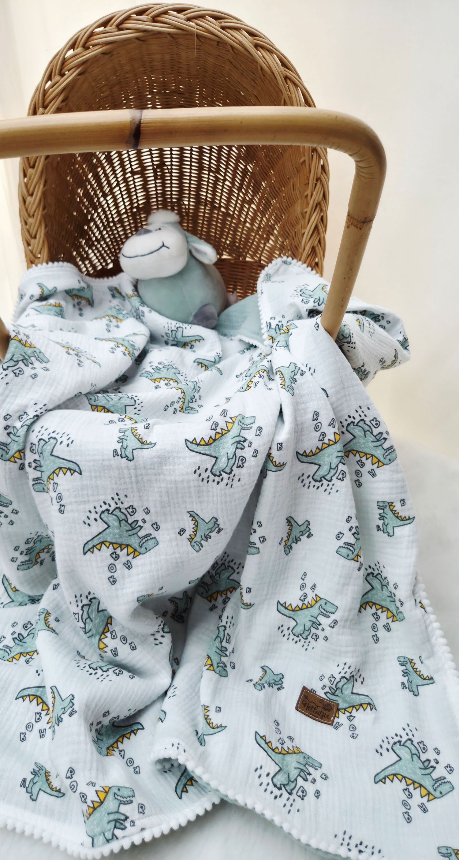 Soft white muslin baby blanket with green dinosaur print and pom-pom trim draped in a wicker basket with a plush lamb toy.