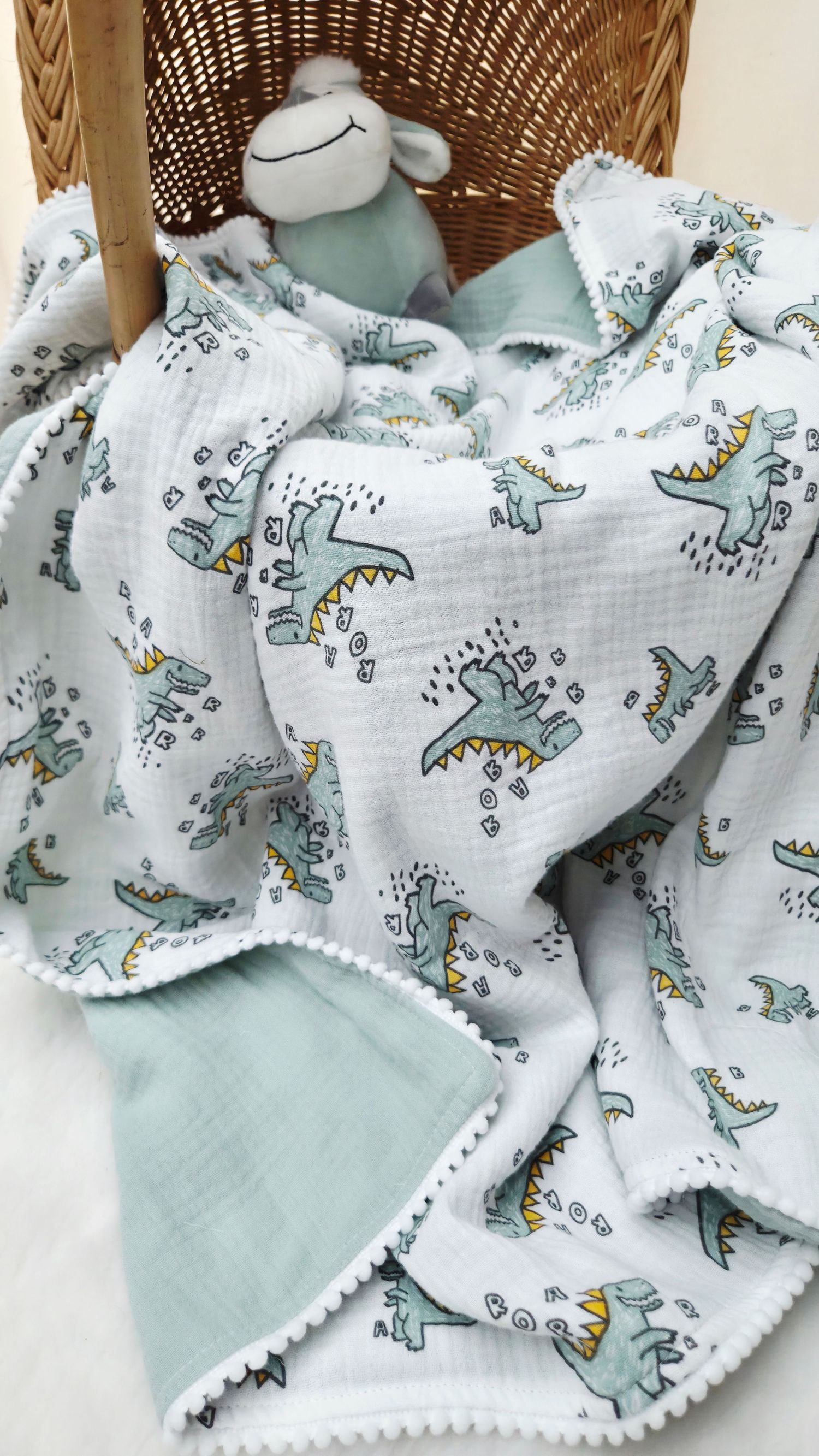 Soft muslin baby blanket with green dinosaur print, sage green backing and pom pom trim, with a plush lamb toy in a wicker basket.
