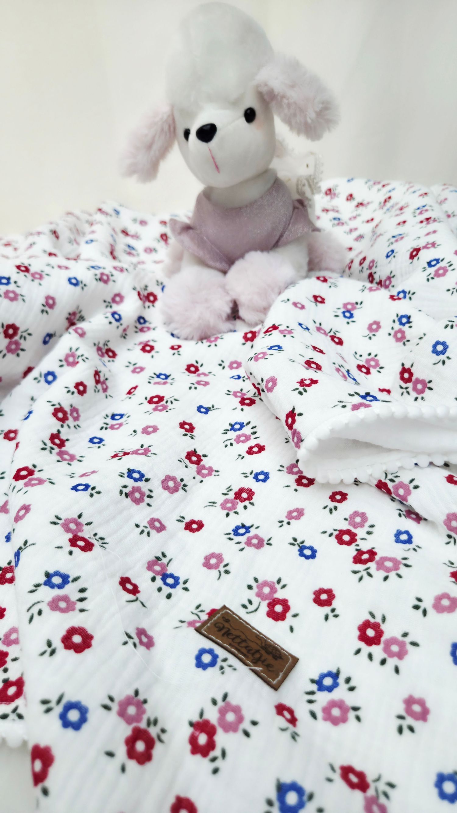 A plush white poodle toy in a lilac sweater sits on a soft white muslin blanket with a red, pink and blue floral print.