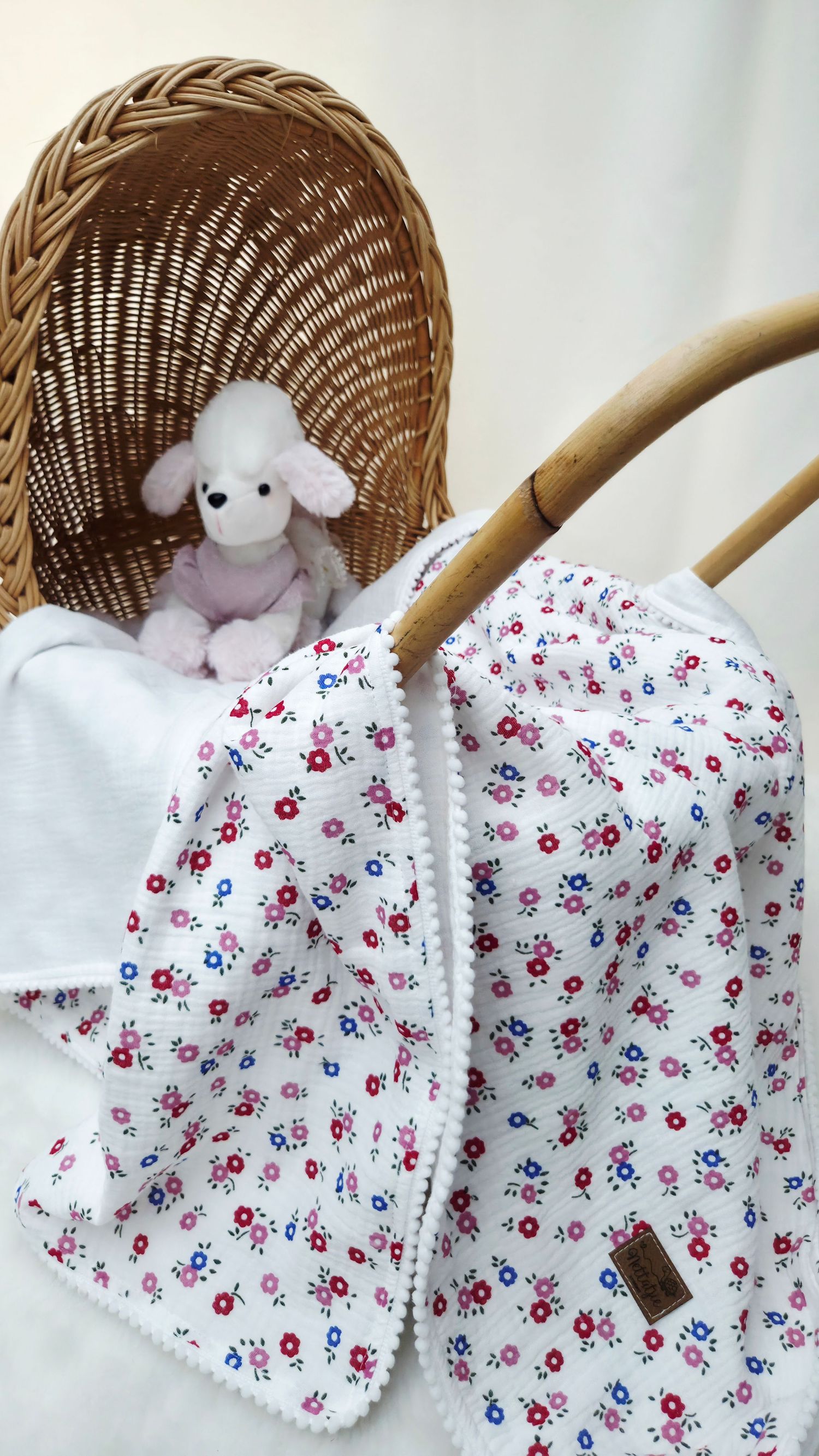 Floral cotton baby blanket with pom-pom trim draped over a wicker pram holding a plush white poodle toy.