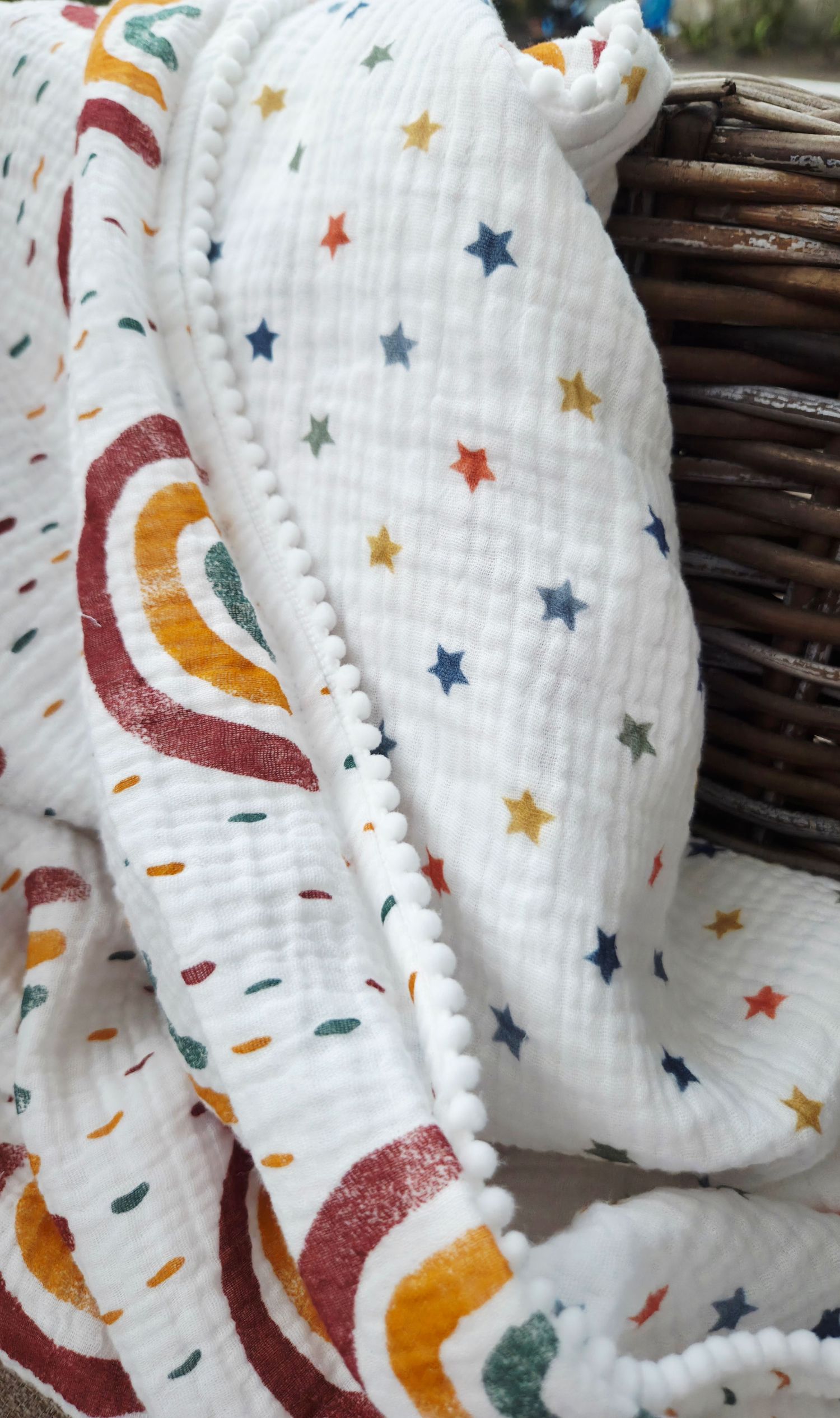 White muslin baby blanket with colorful stars, rainbow print and pom pom trim, draped over a woven wicker basket.