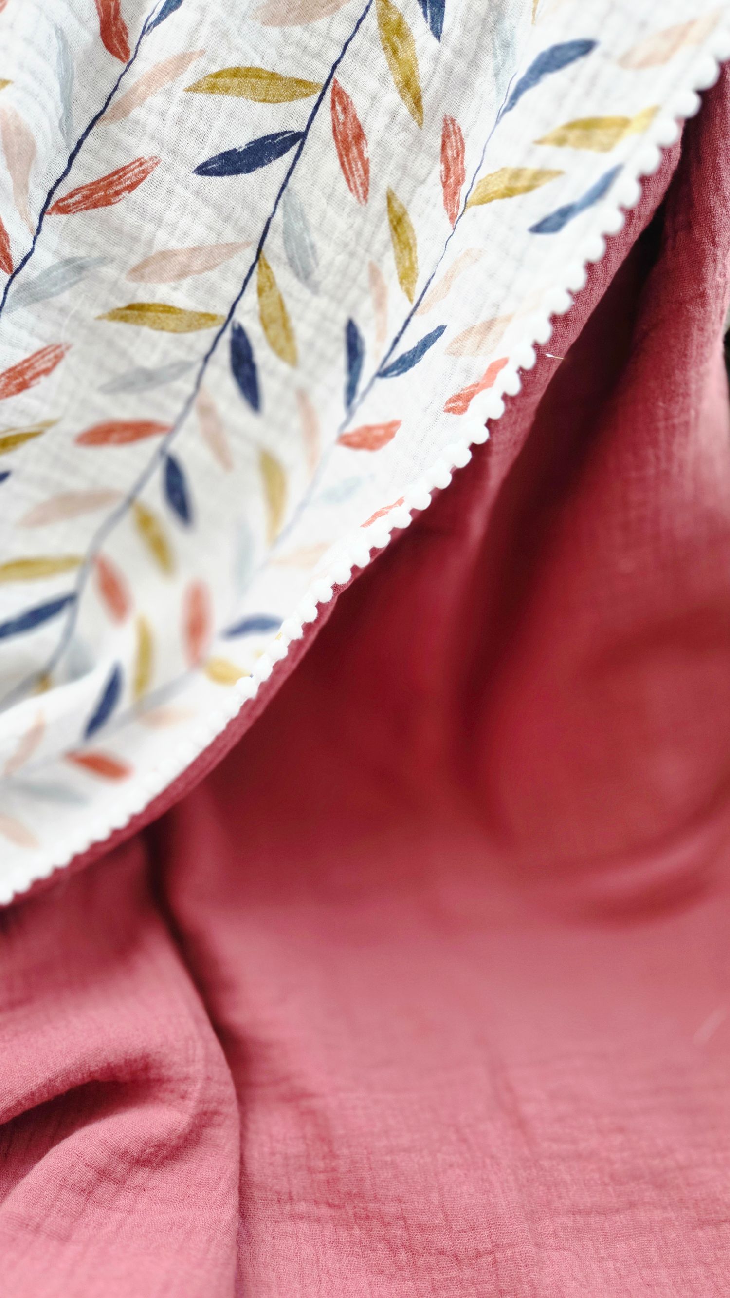 Double gauze muslin blanket with a leaf print in navy, mustard and coral on cream, edged with white pom-pom trim over dusty rose cotton.