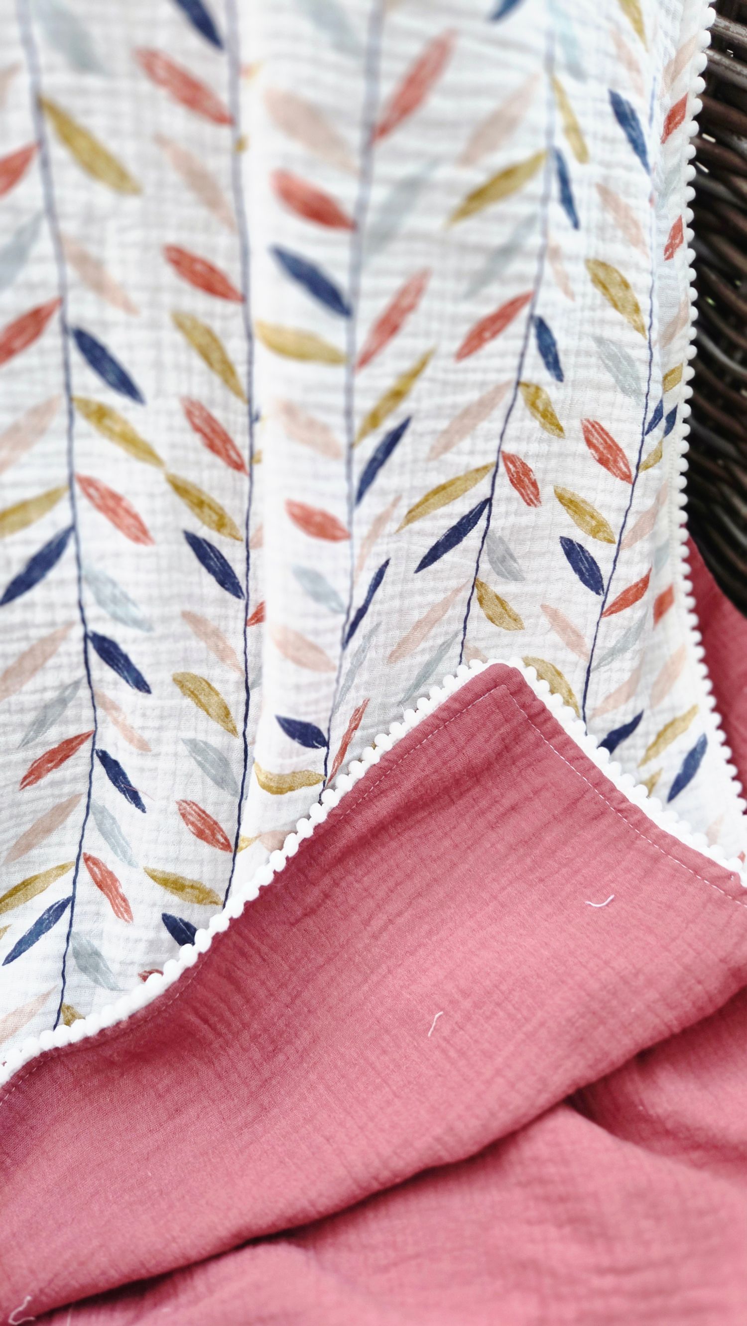 Reversible muslin baby blanket with a leaf print in navy, rust and mustard on white, a white pom pom trim and a dusty rose backing.