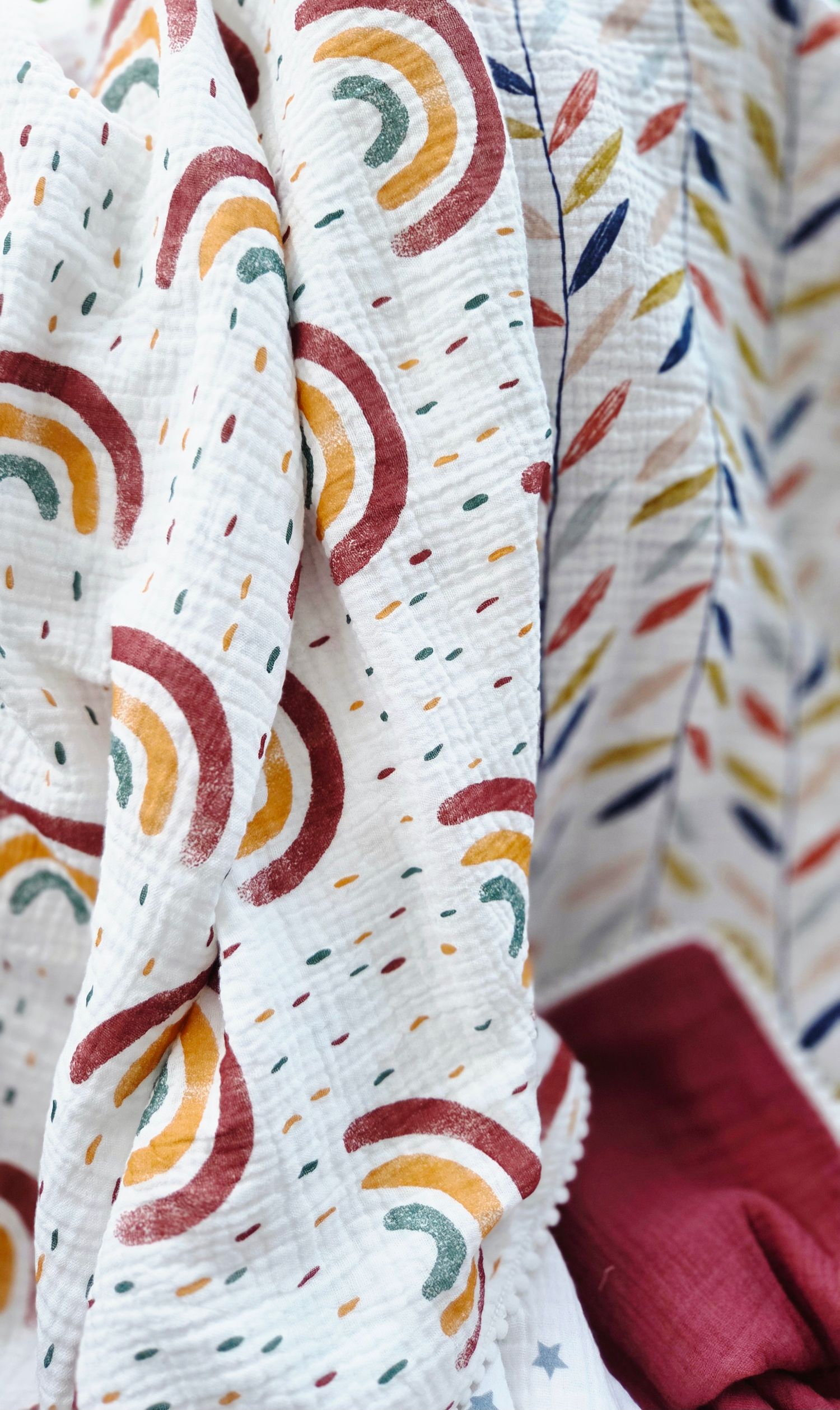 Muslin baby blanket with rainbow print and colorful leaf pattern on the reverse, finished with a burgundy border and white trim.