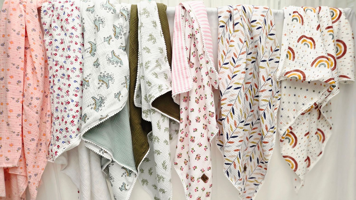 Pastel baby muslin swaddle blankets with floral, animal, striped and rainbow prints hanging in a row against a white wall.
