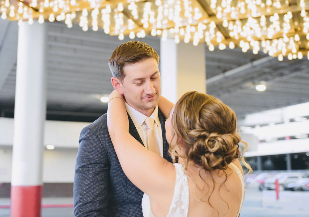 Groom smiles tenderly as bride places hands on his shoulders under warm string lights at wedding.