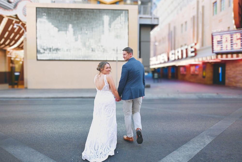 Bride in lace gown and groom walking hand in hand on Las Vegas Fremont Street at sunset.