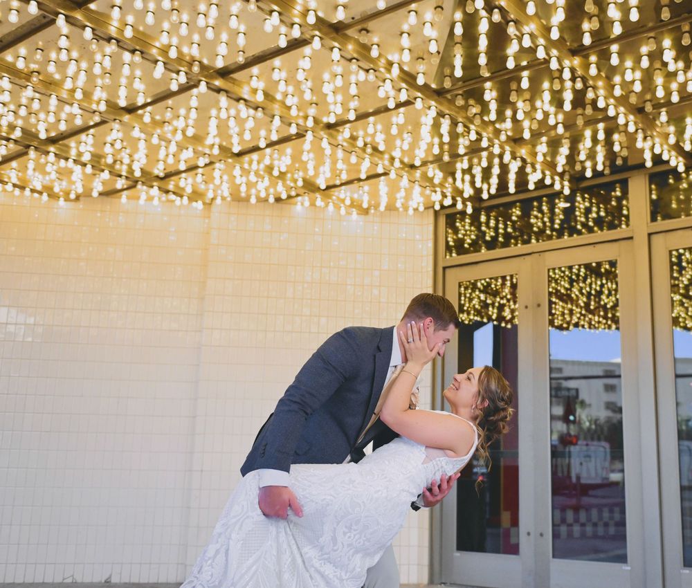 Bride and groom share a romantic dip kiss under sparkling string lights at their wedding venue.