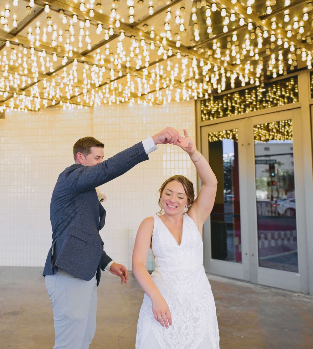 Bride in white gown being twirled by groom in blue suit under sparkling lights at wedding reception.