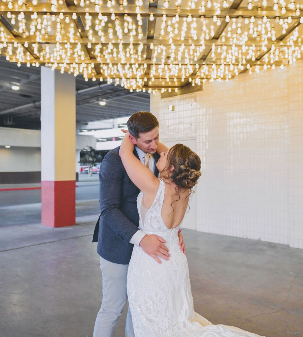 Bride and groom share romantic embrace under string lights in stylish urban venue setting.