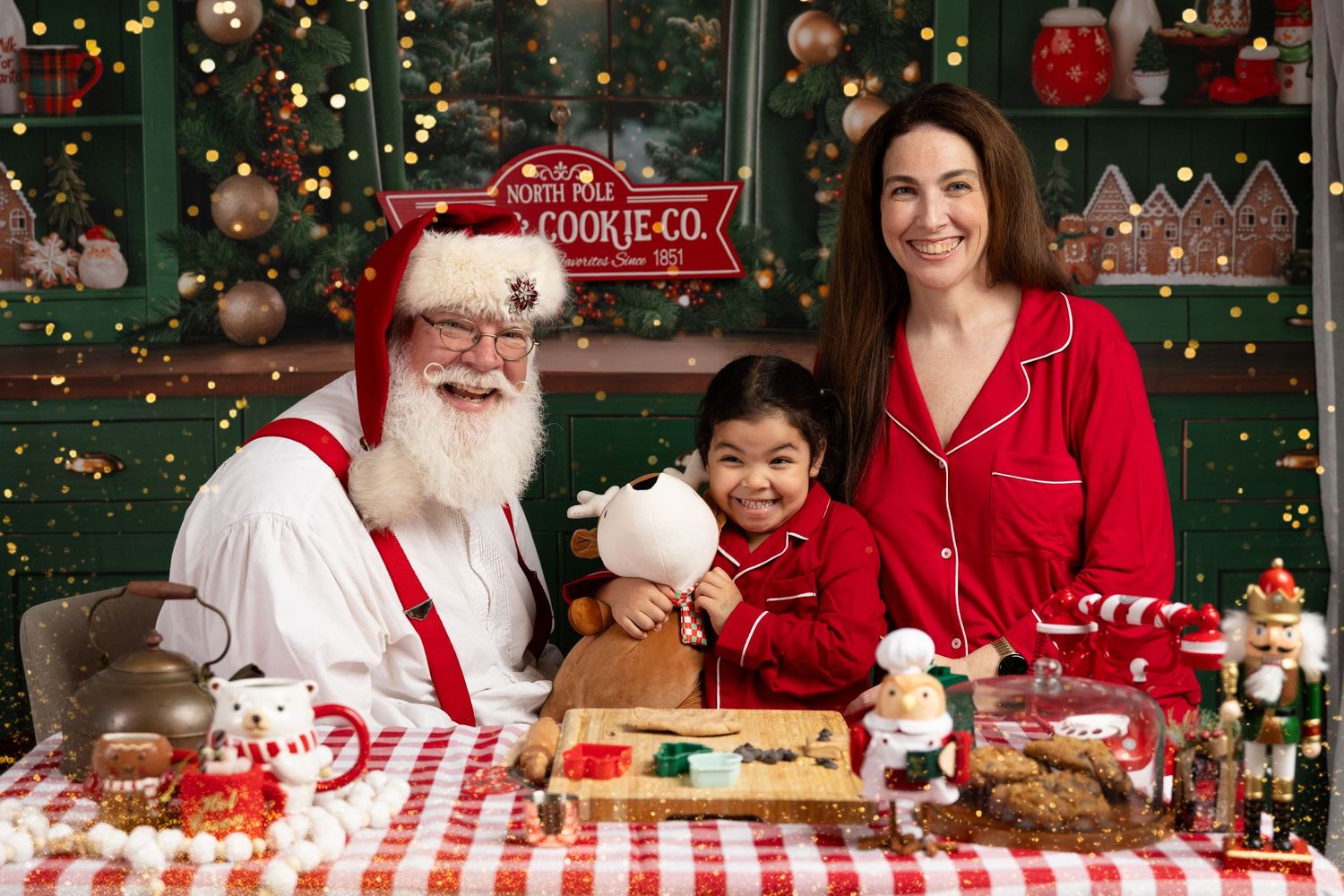 Santa Claus poses with a woman and child at a festive Christmas cookie decorating table with holiday decorations.
