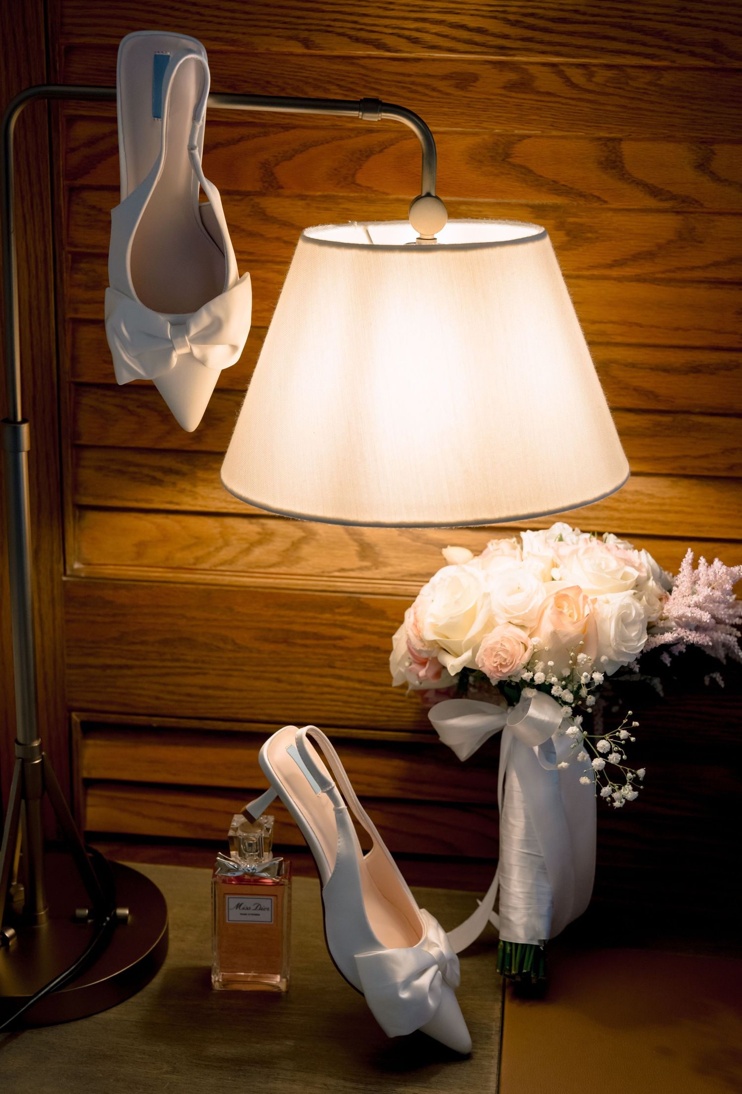 White bridal heels with bow accents displayed near a glowing lamp and fresh rose bouquet on a wooden surface.