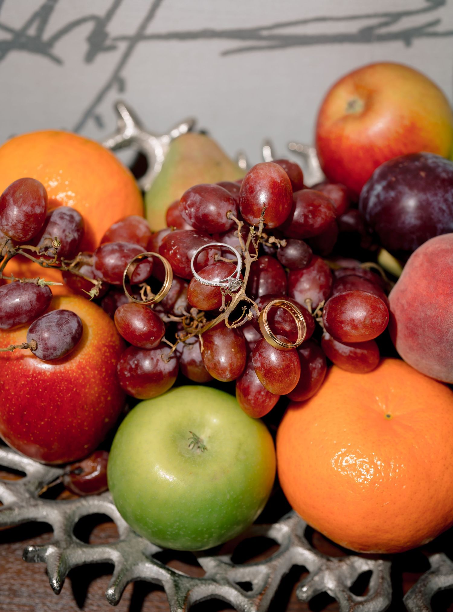 A decorative silver bowl overflowing with fresh fruit including red grapes, oranges, apples, pears, and a peach.