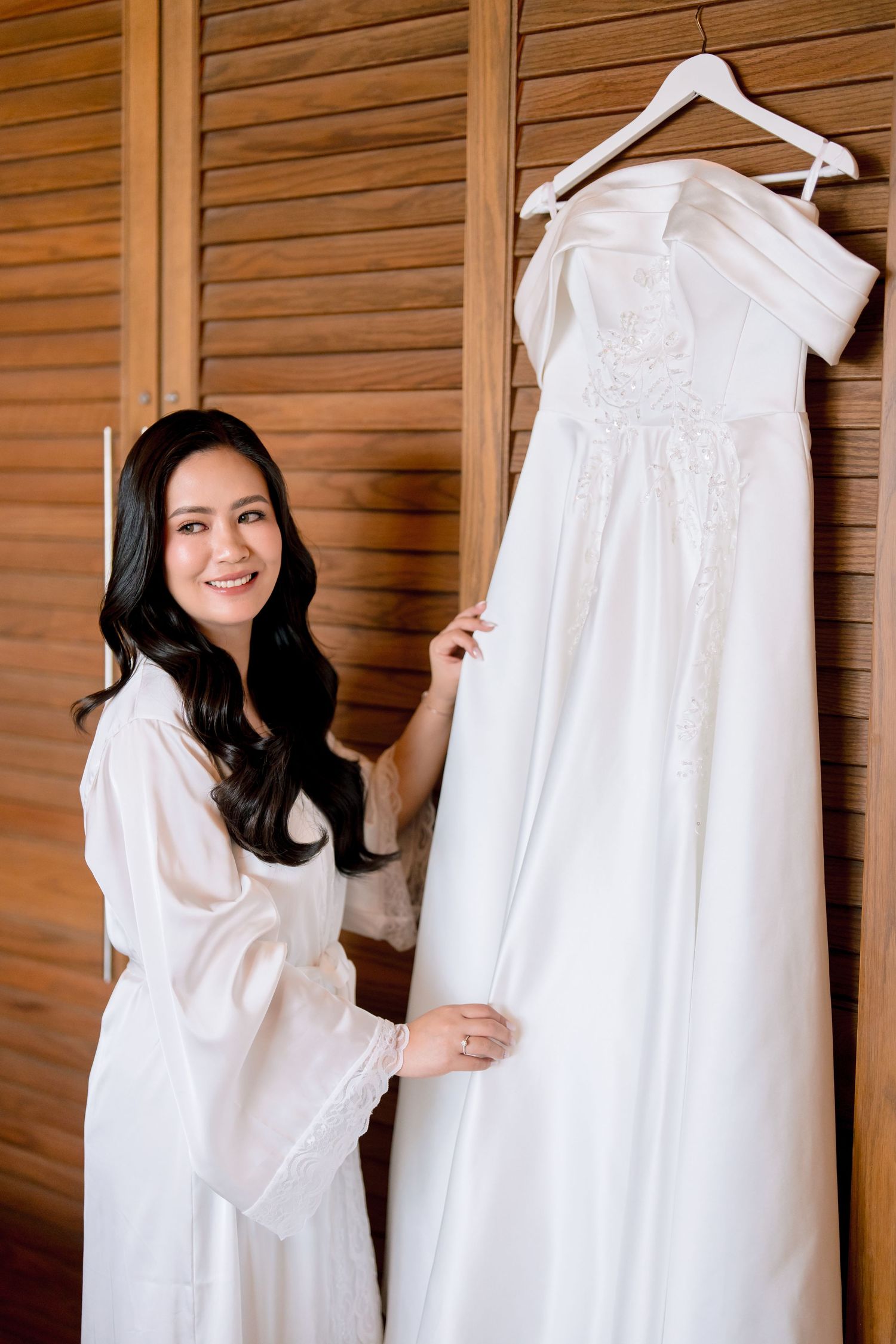 Smiling bride in white robe admiring her elegant strapless wedding gown hanging on wooden door.