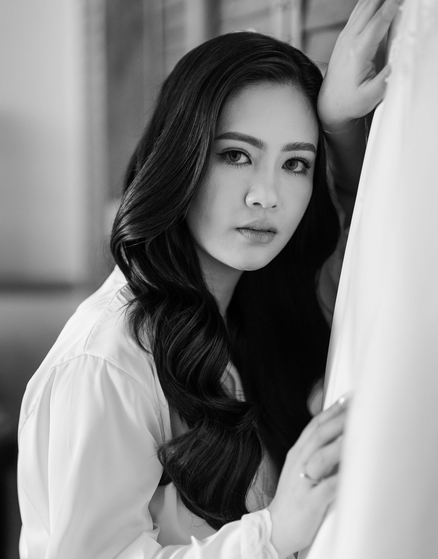 Black and white portrait of a young Asian woman with long wavy hair leaning against a white surface.