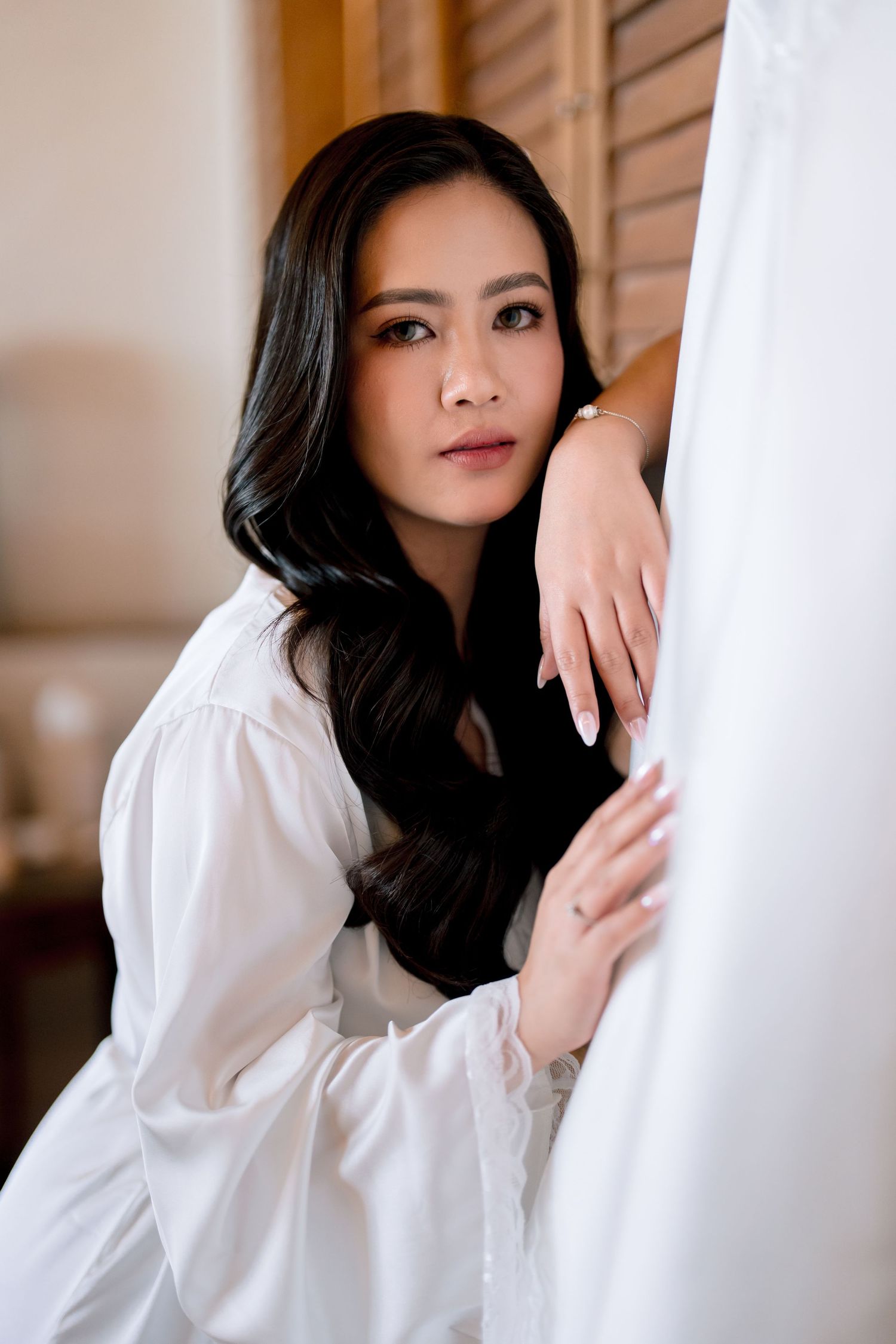 A young Asian woman in a white shirt leans gracefully against a white curtain, gazing softly at the camera.