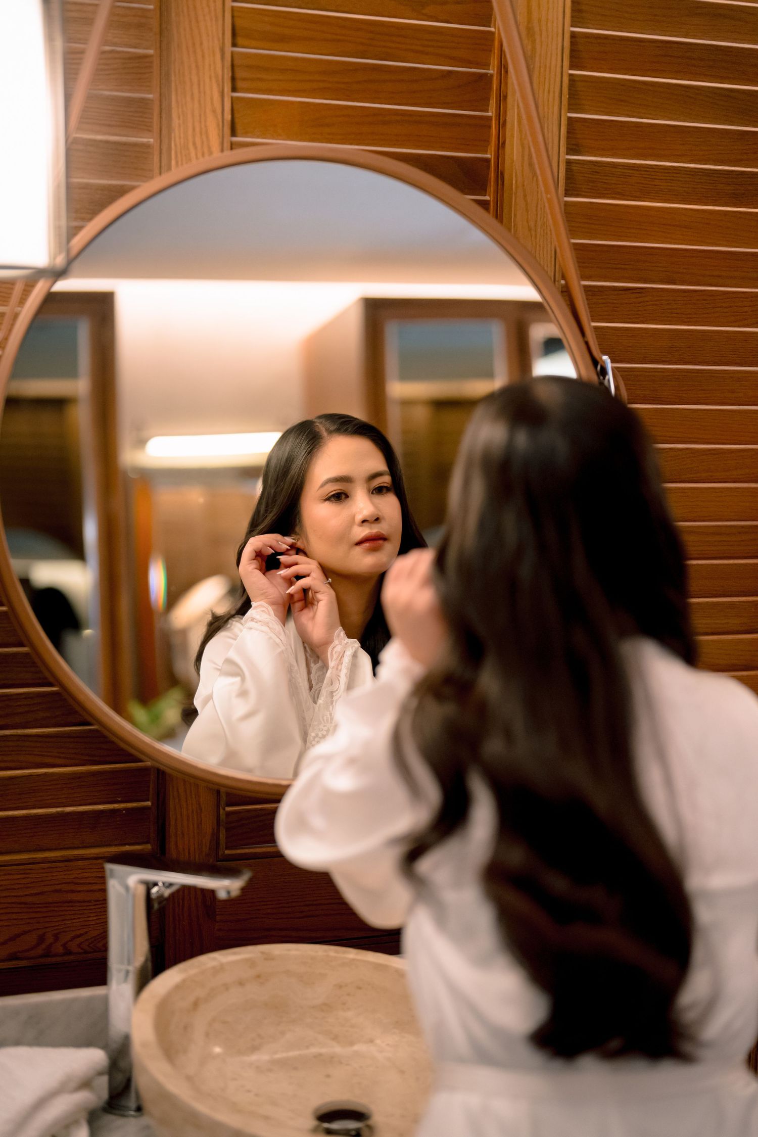 Young Asian woman in white robe putting on earrings while looking at round mirror in elegant bathroom.