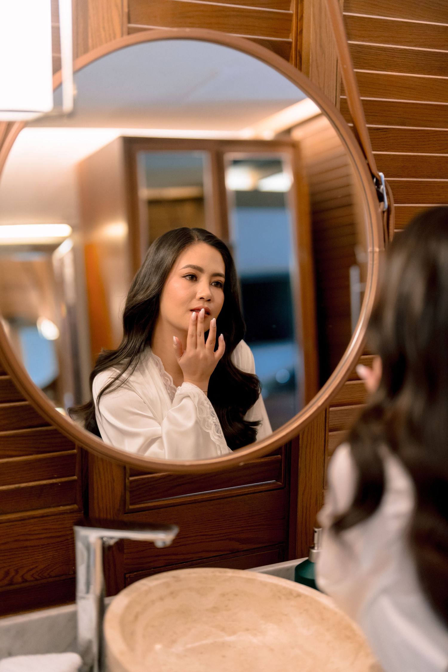 Young Asian woman in white shirt applying lip care while looking at her reflection in a round bathroom mirror.