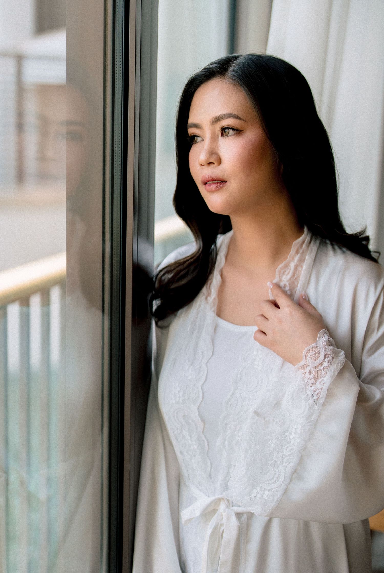 A woman in white lace robe gazes thoughtfully out a window, her dark hair flowing over her shoulders.