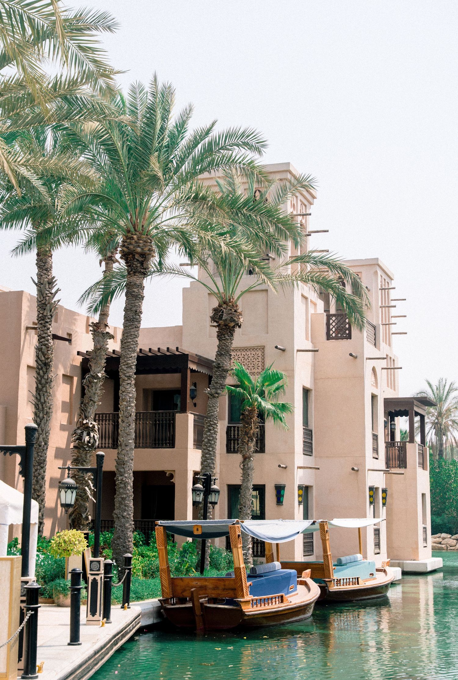 Traditional Arabic boats dock along a serene waterway beside a luxury Middle Eastern resort with palm trees.