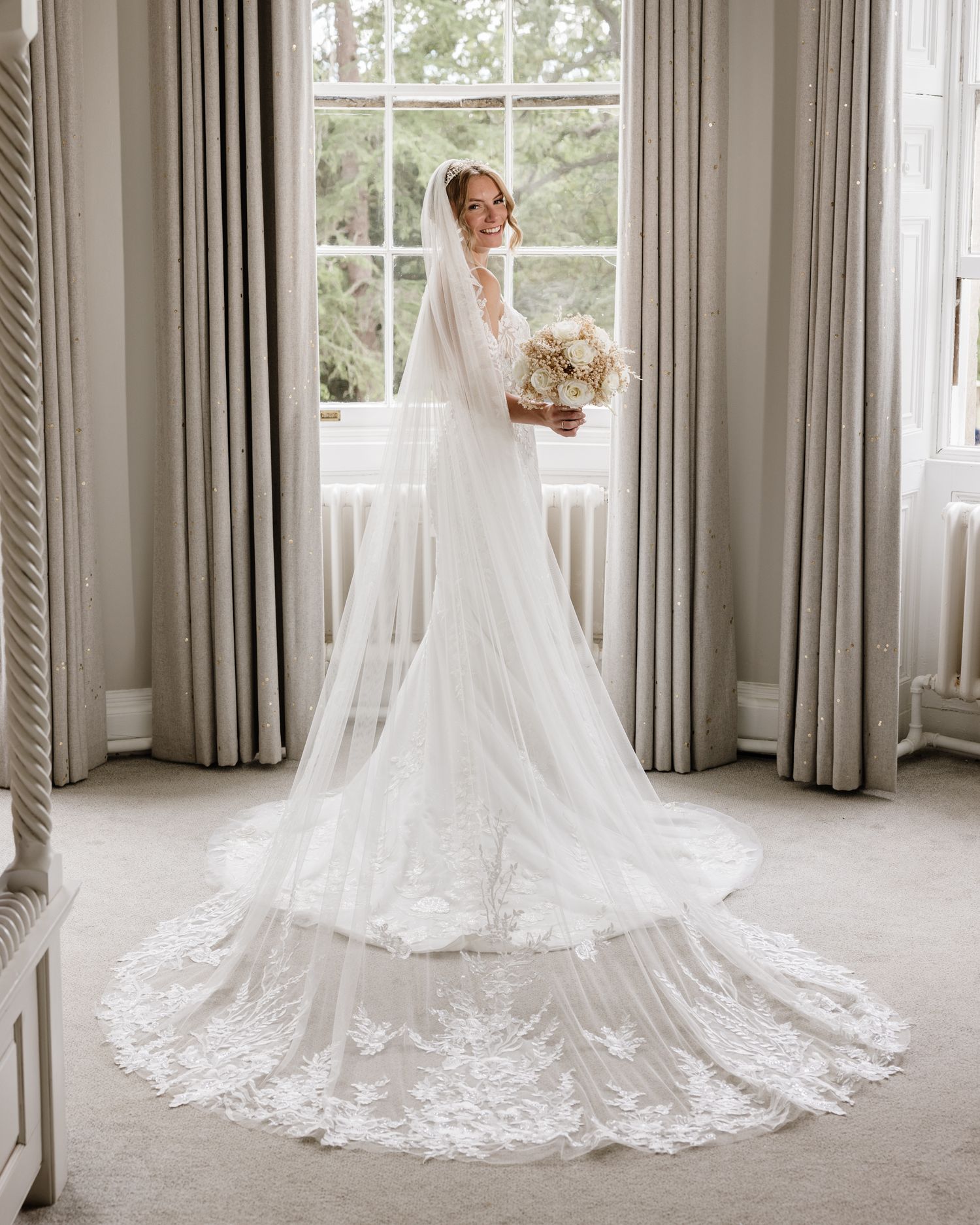 Bride in elegant long-sleeve white wedding gown with lace cathedral veil, holding bouquet by a bright window.