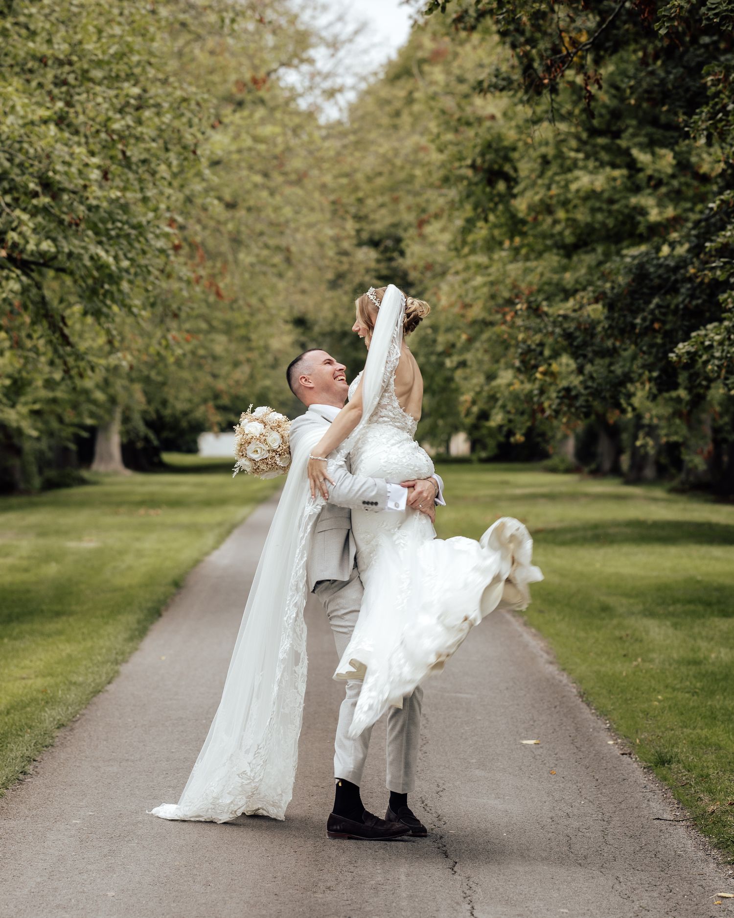 Groom lifting bride in white gown and veil on tree-lined path during romantic outdoor wedding photo.