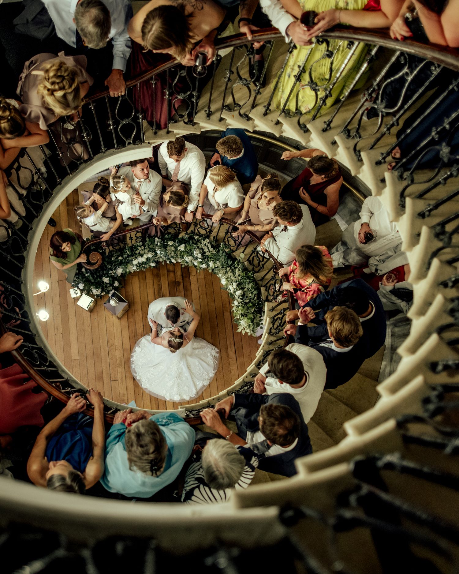 Aerial view of a bride in a white gown at the center of an elegant spiral staircase surrounded by wedding guests.