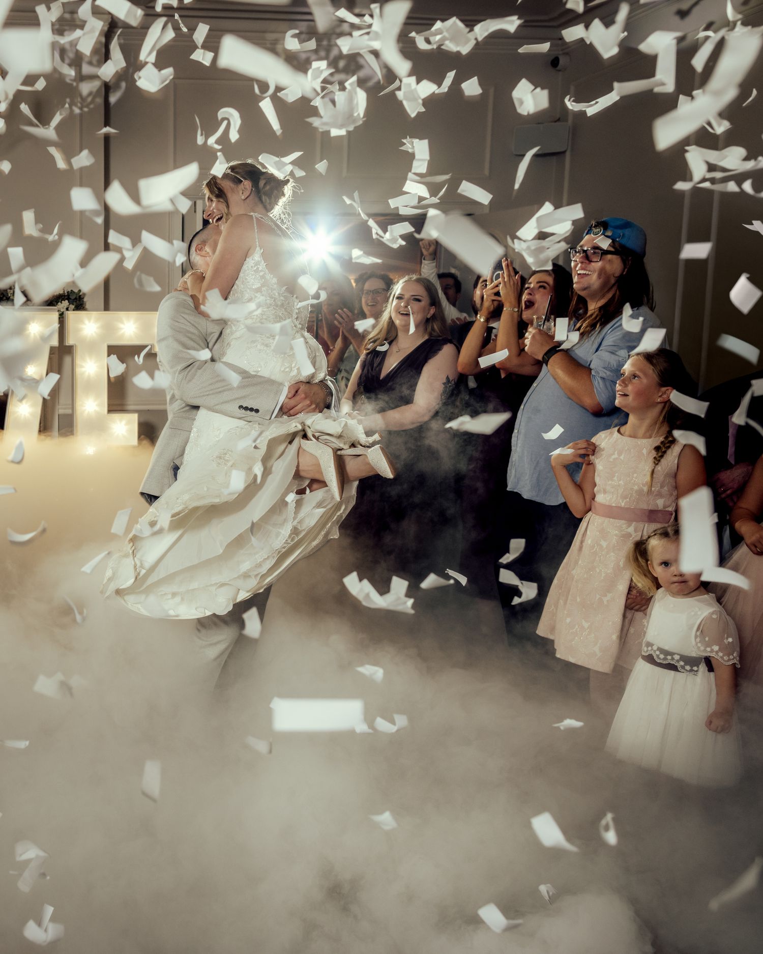 Bride and groom celebrate with confetti and fog at their wedding reception surrounded by guests and a flower girl.