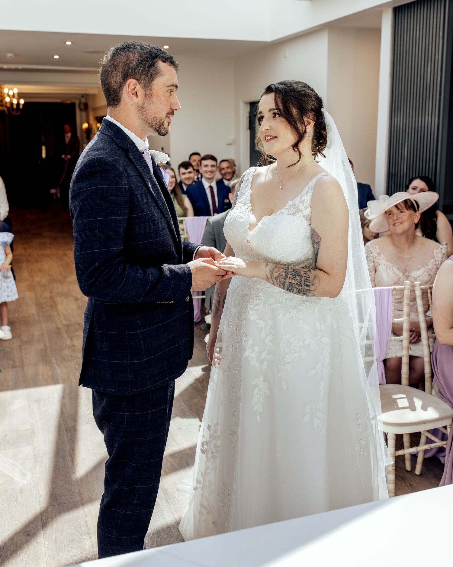 Bride and groom exchange rings during indoor wedding ceremony, guests seated in background.