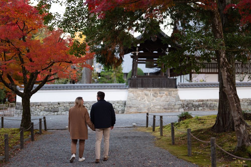 Couple holding hands walking toward a traditional Japanese temple bell tower surrounded by vibrant autumn foliage.