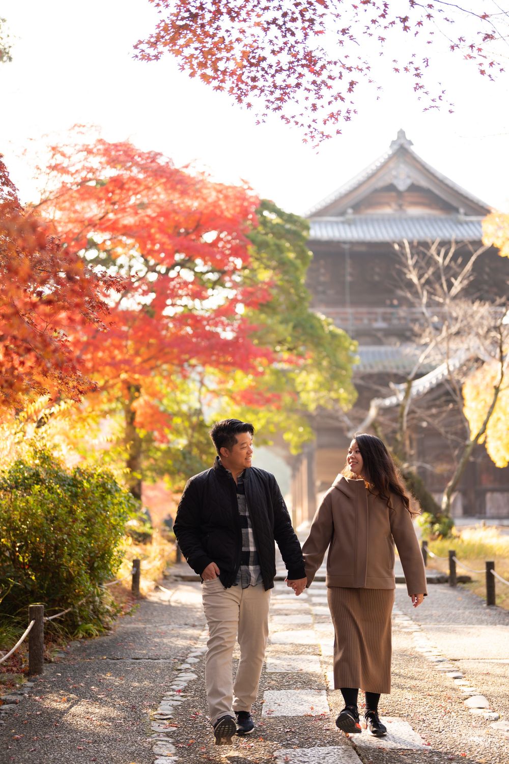 Couple holding hands walking on a stone path surrounded by vibrant autumn foliage near a traditional Japanese temple.