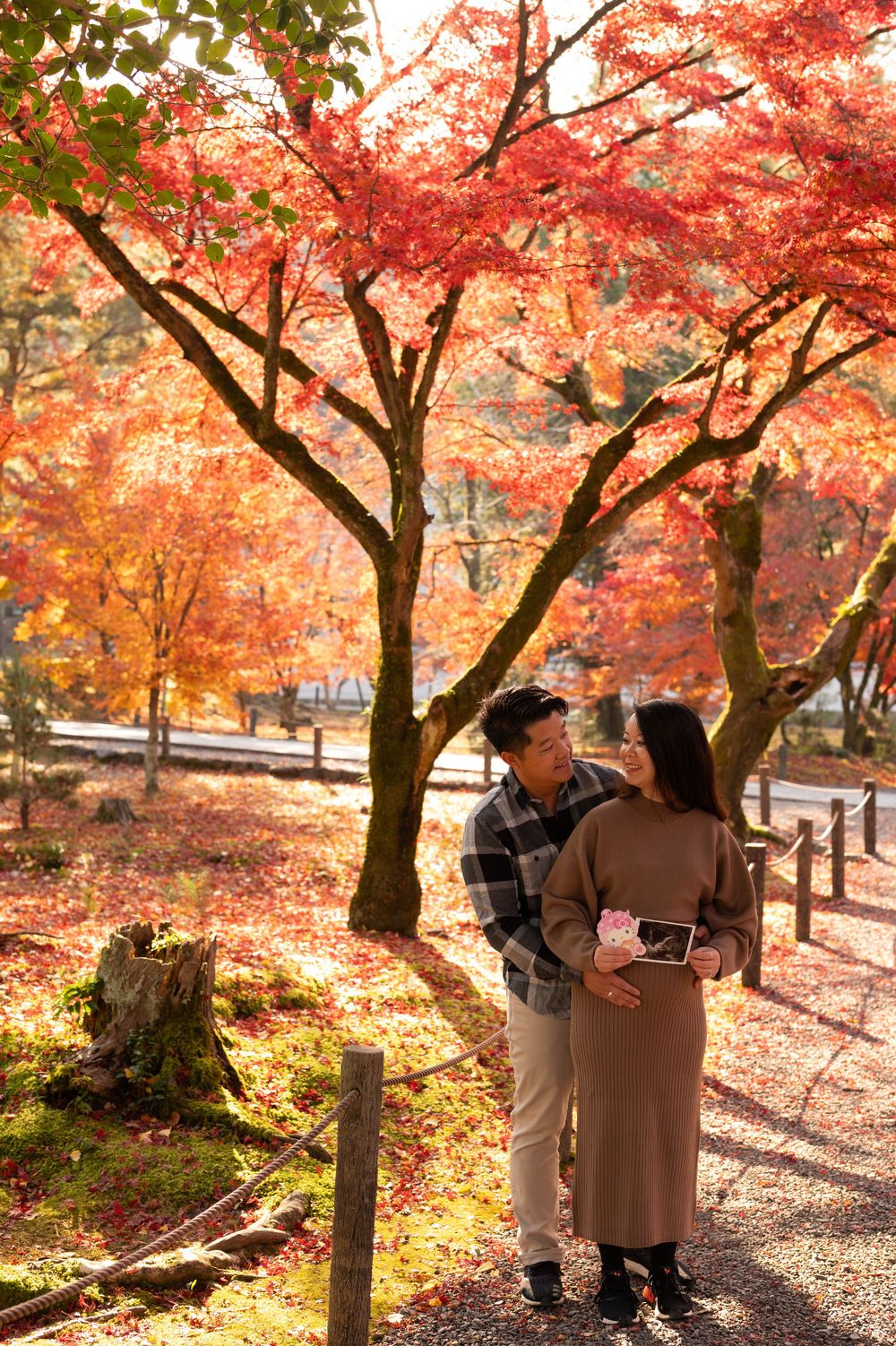 Couple sharing a tender moment under vibrant red maple trees in autumn park, woman holding ultrasound photo.