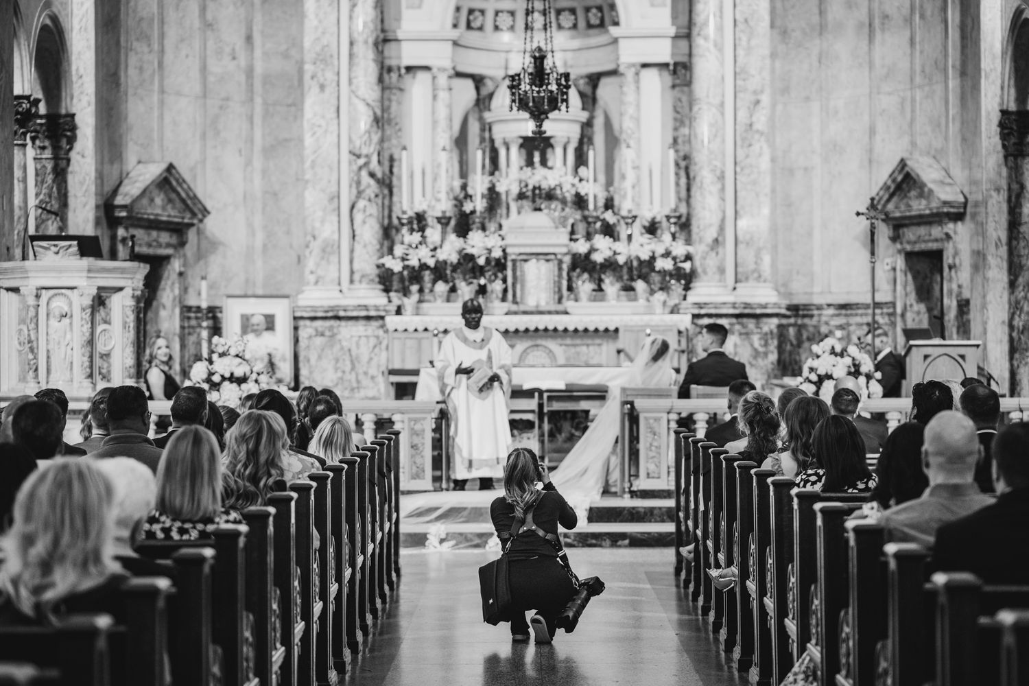 Black and white photo of a wedding ceremony in a grand church as guests fill the pews and a photographer crouches in the aisle.