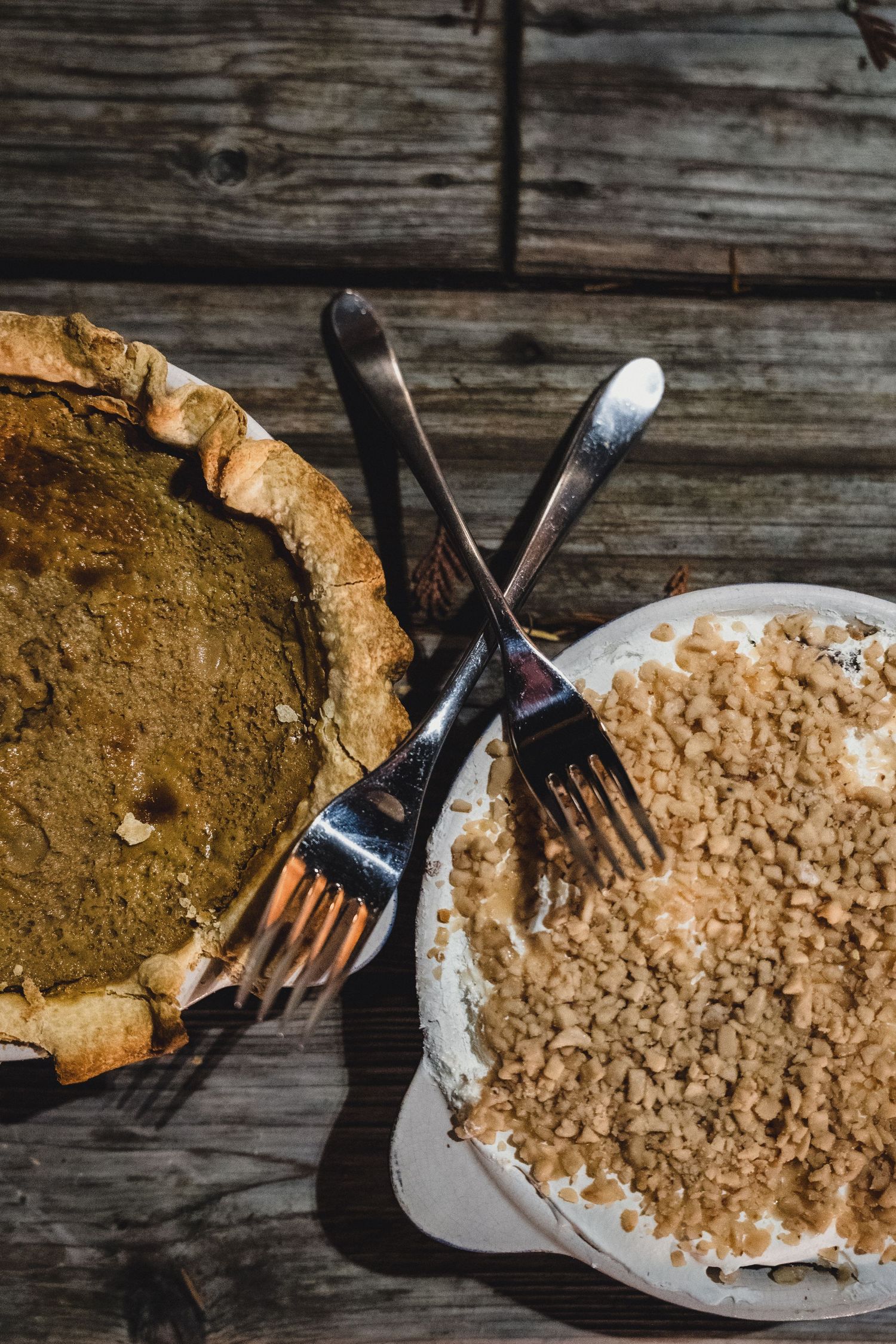 Two forks rest across a golden pumpkin pie and a crumble-topped dessert on a rustic wooden table.