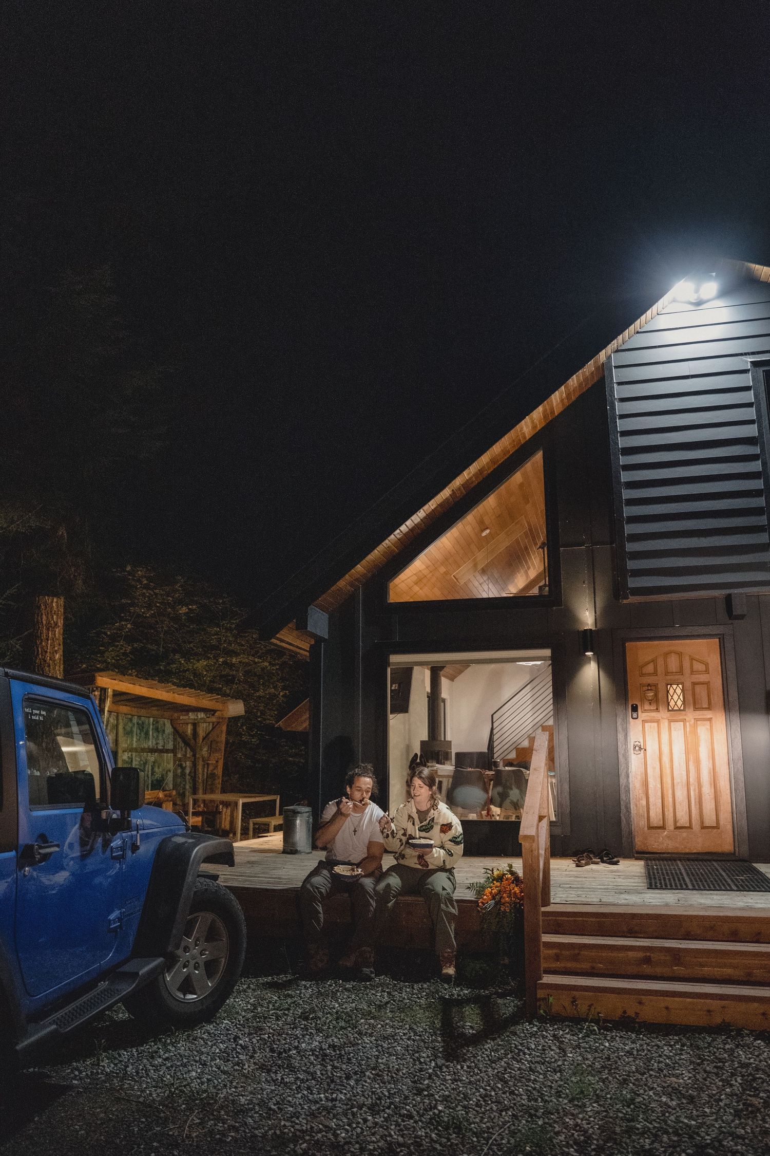 Two people sit on the porch of a modern dark-sided cabin at night, with a blue Jeep parked nearby on gravel under warm glowing lights.