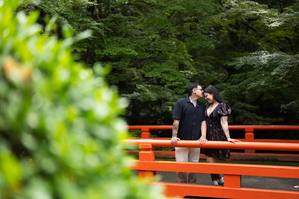 Couple kissing on a red wooden bridge surrounded by lush green trees in a peaceful garden during an engagement photo session.