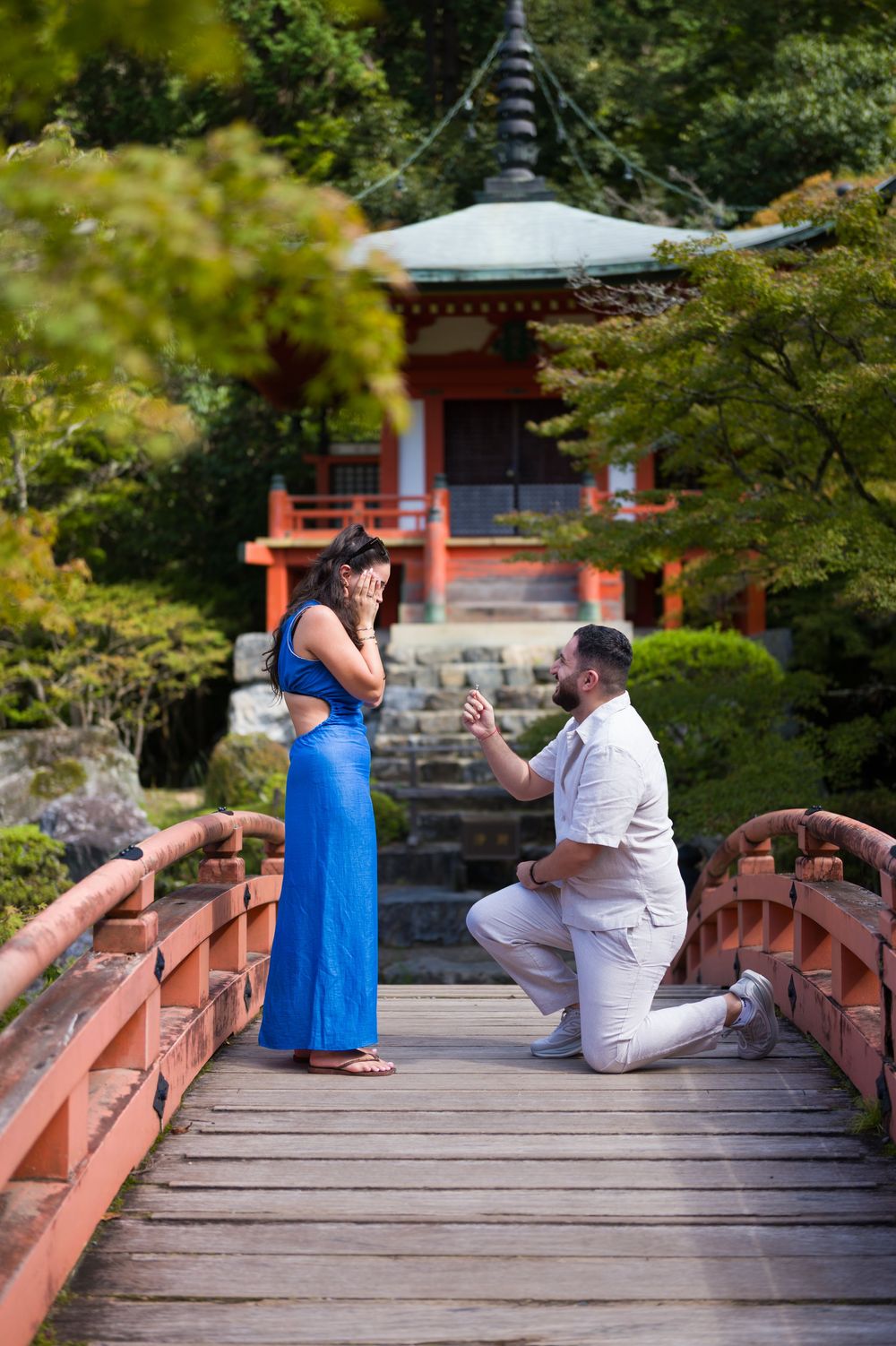 Man kneels on one knee proposing to a woman in a blue dress on a red wooden bridge in front of a Japanese temple.