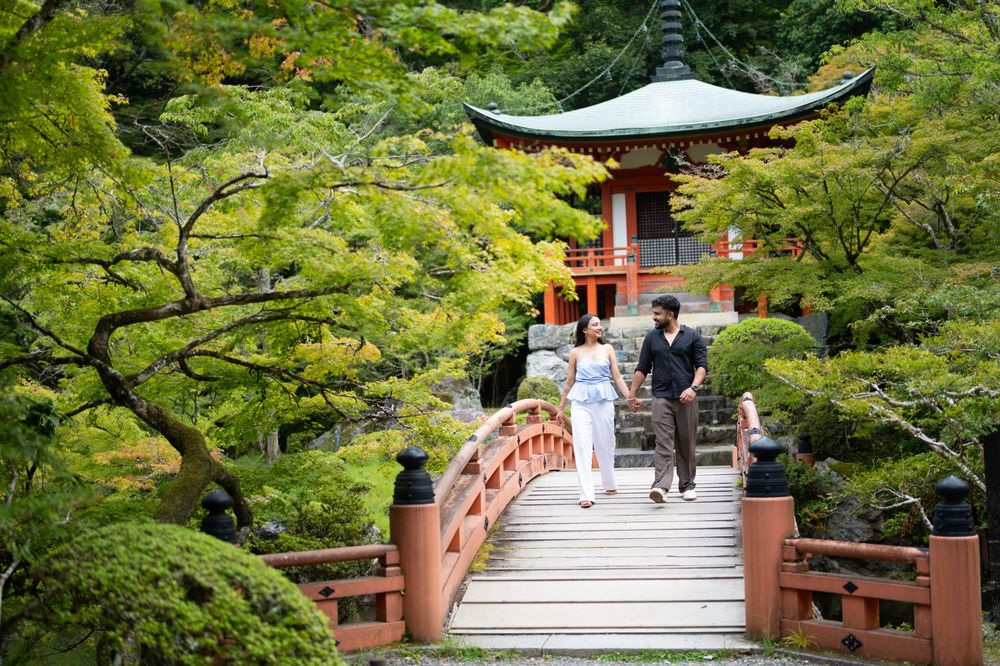 A couple walks across a red wooden arched bridge toward a traditional Japanese pagoda surrounded by lush green maple trees.
