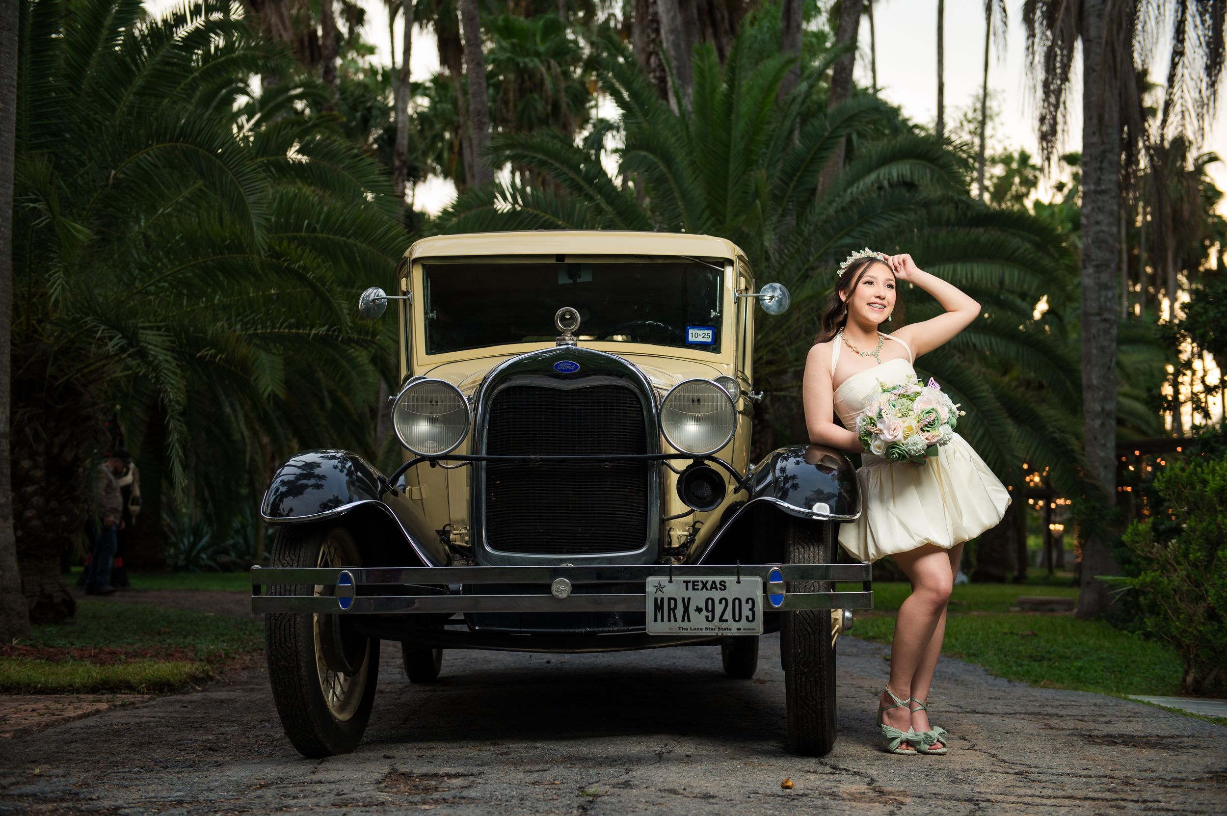 Bride in white dress poses beside a vintage classic car surrounded by lush palm trees in an elegant outdoor setting.