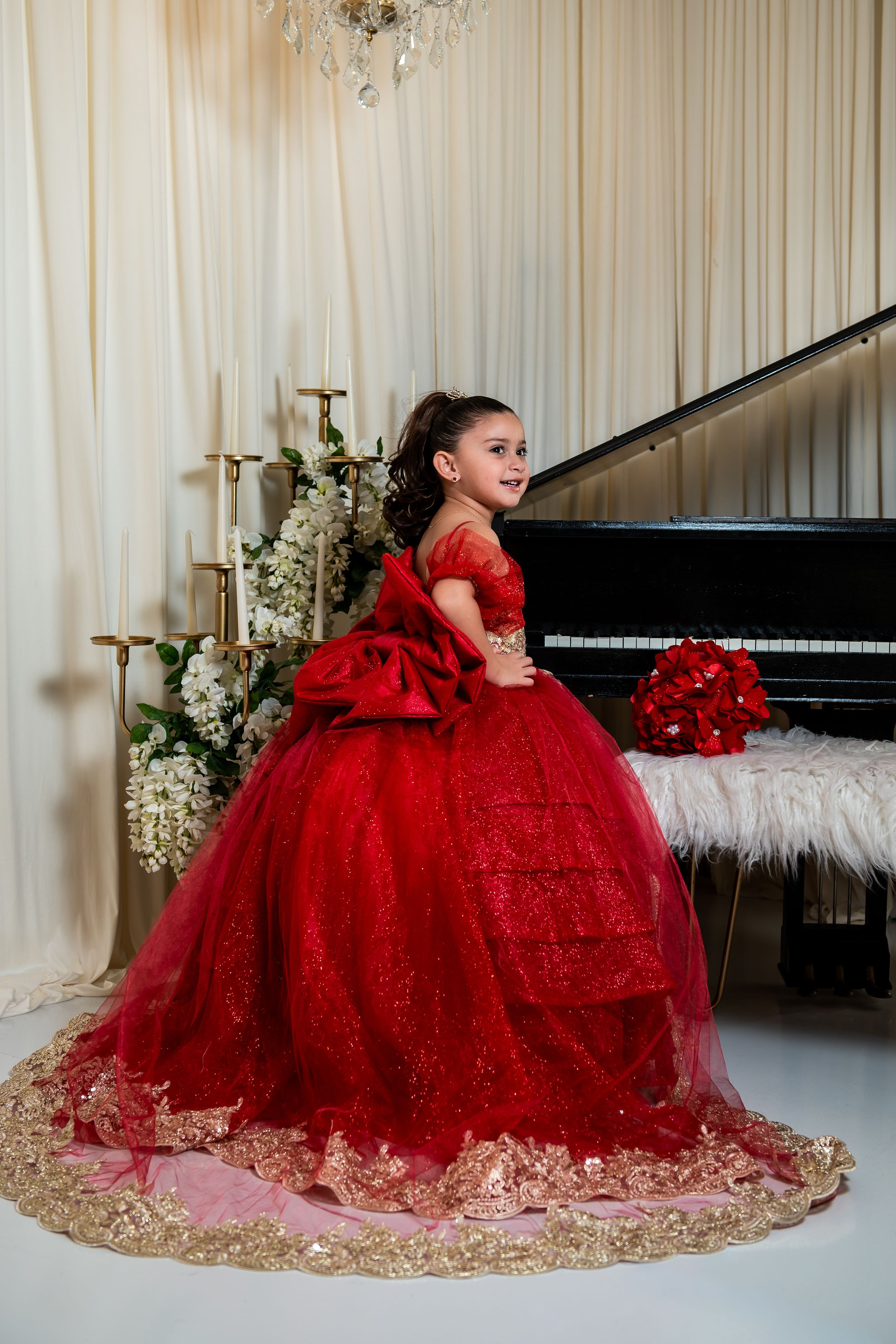 Young girl in elegant red ballgown with gold trim poses beside a grand piano in a decorated studio.