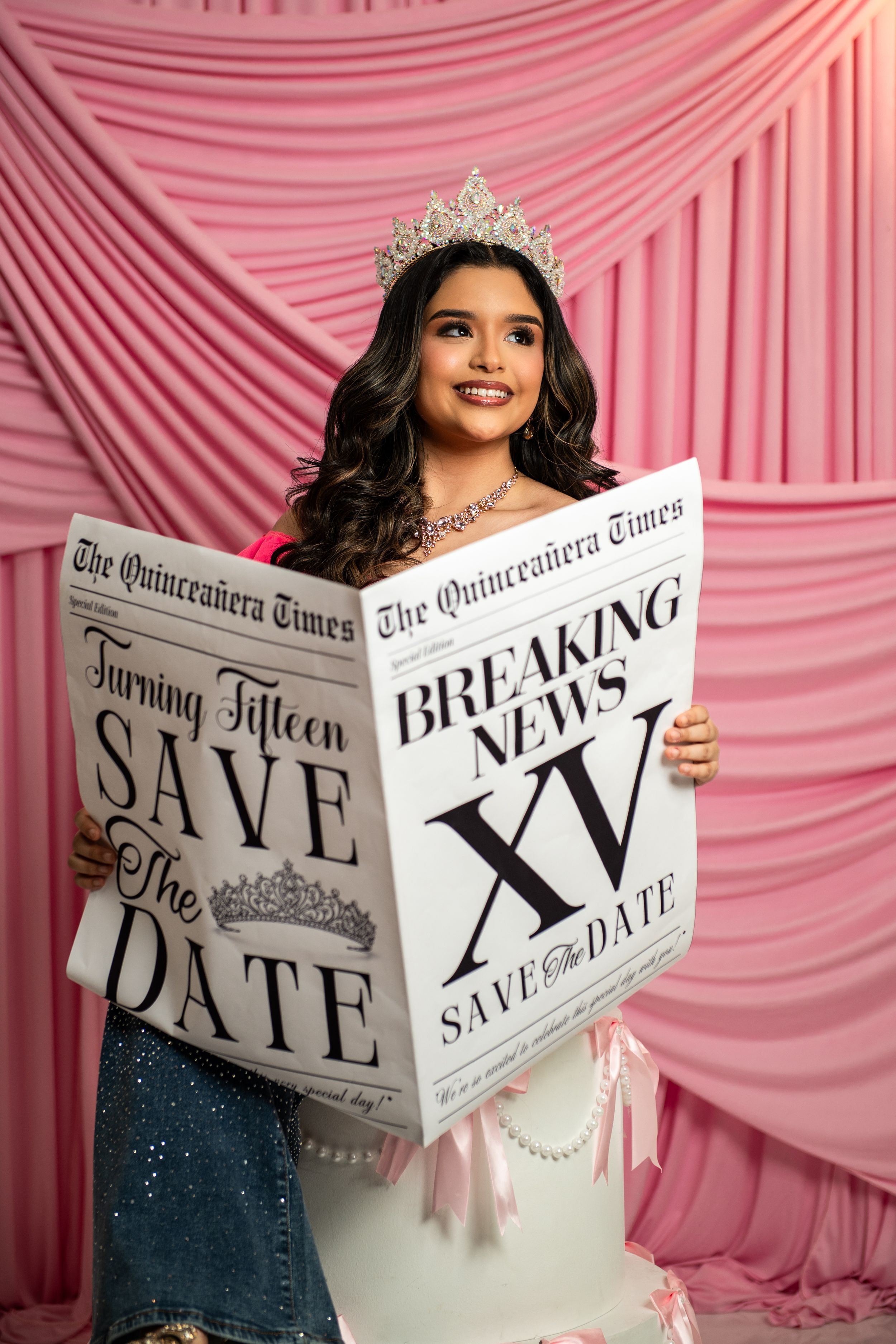 Young girl in tiara holds Quinceanera Times Save the Date newspaper prop against pink backdrop.