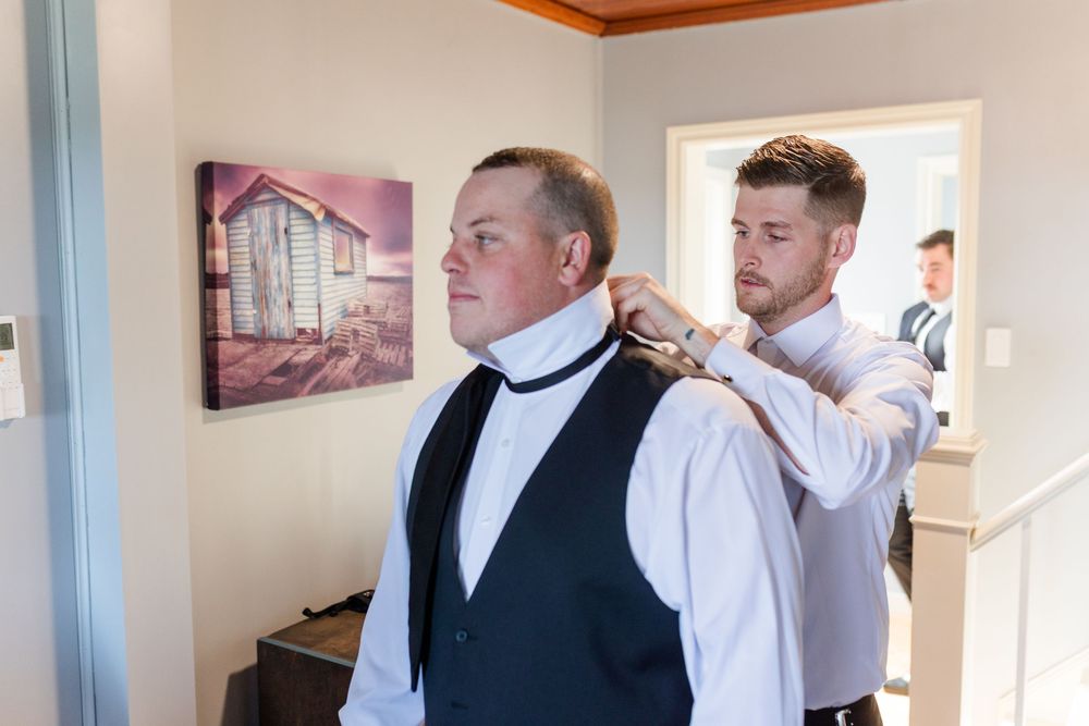 Groomsman adjusting the collar of a groom in a black vest and white shirt before a wedding ceremony.