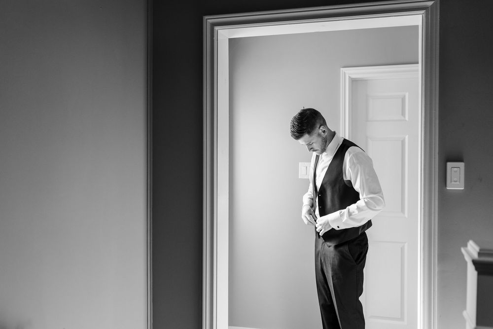Black and white photo of a groom adjusting his vest in a doorway while preparing for his wedding day.