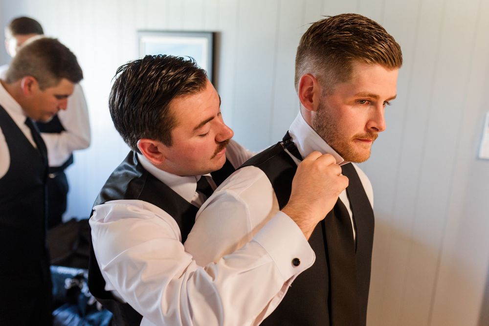 Groomsman adjusting the tie of the groom while another groomsman dresses in the background.