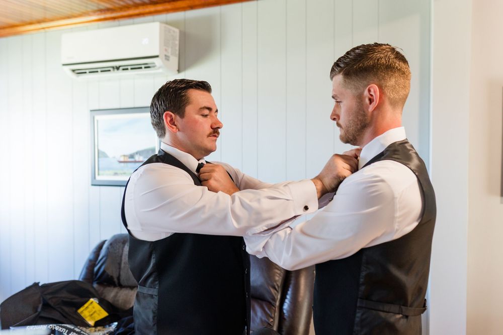 Two groomsmen in vests helping each other adjust ties while getting ready for a wedding.