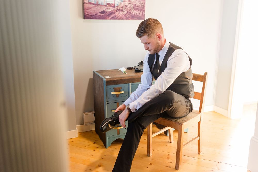 Groom in vest and tie sitting on wooden chair putting on dress shoes before wedding ceremony.