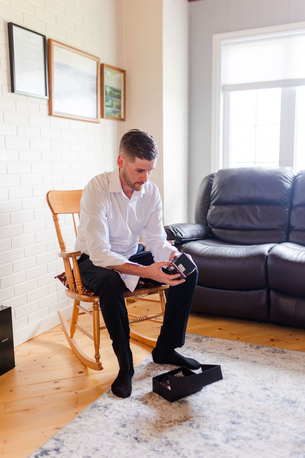 Groom sitting in a wooden rocking chair putting on dress shoes before his wedding ceremony.